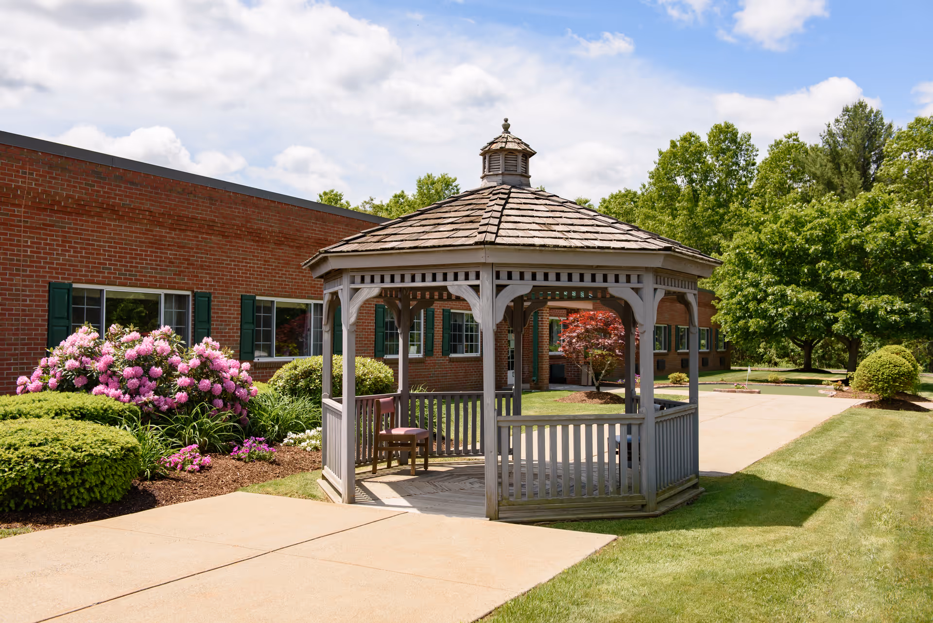 A wooden gazebo with a shingled roof is situated on a paved walkway surrounded by well-maintained green lawns, bushes, and blooming pink flowers. Behind the gazebo is a red brick building with green window shutters, and trees are visible in the background under a partly cloudy sky.