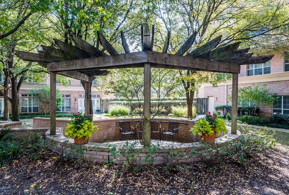 Brick courtyard with a wooden pergola, potted plants, and metal chairs surrounded by trees and the building.