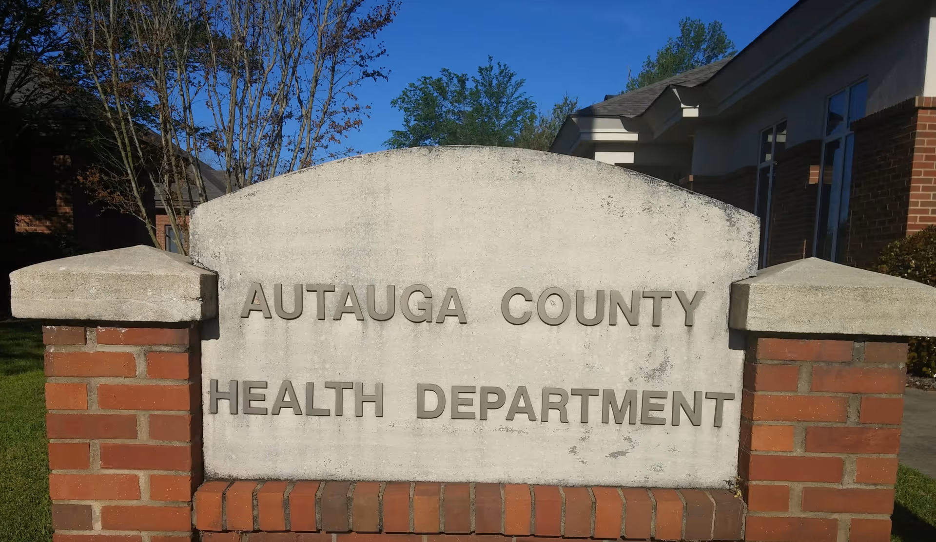 Concrete sign reading 'AUTAUGA COUNTY HEALTH DEPARTMENT' between brick pillars in front of a building.