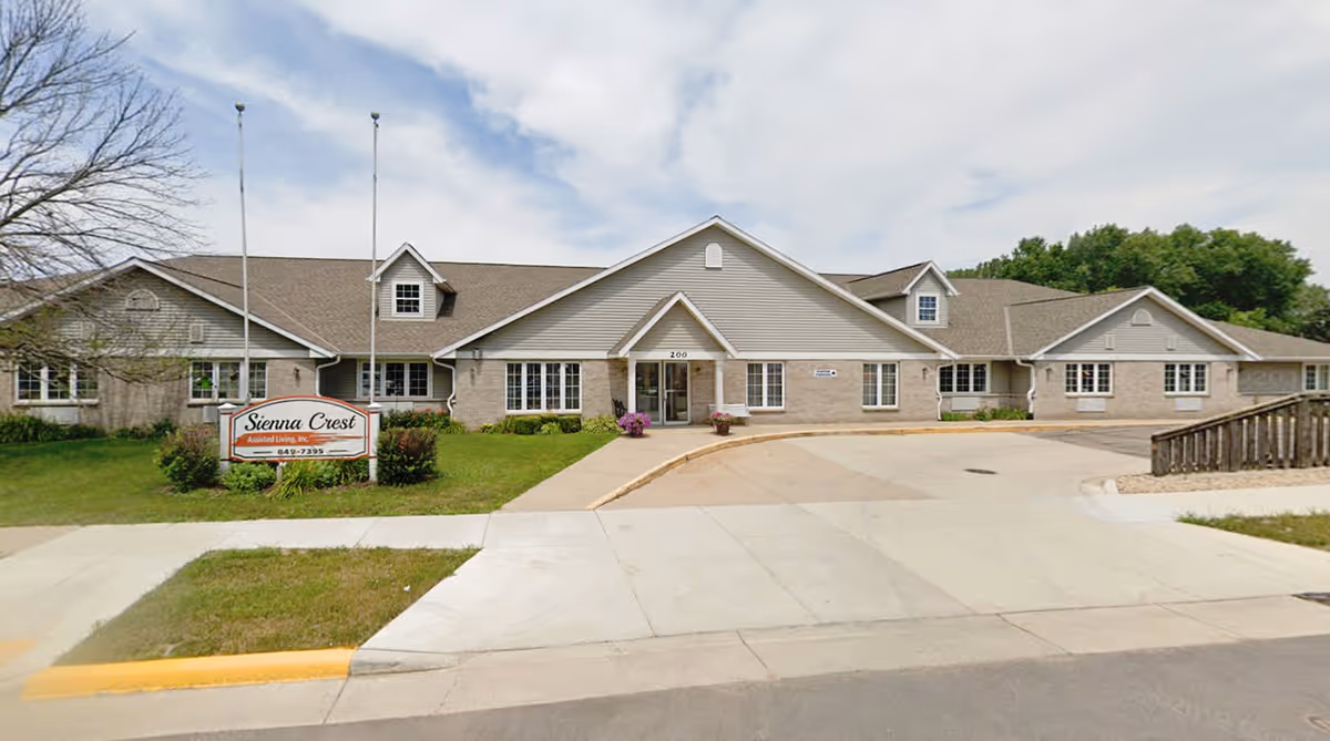 Front exterior view of Sienna Crest Assisted Living, Inc., a single-story building with a light gray roof and beige siding. There is a driveway leading to the entrance, a sign with the facility name on the lawn, and two flagpoles without flags. Trees and greenery surround the building under a partly cloudy sky.