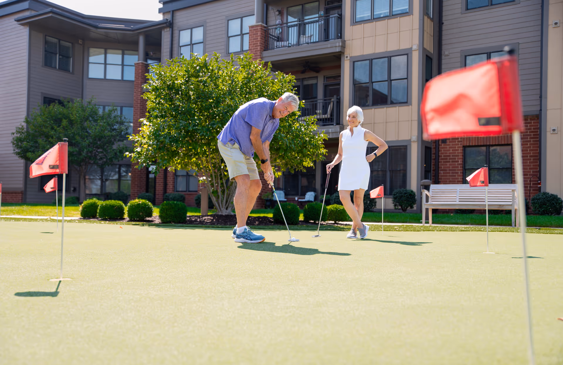 Two seniors play golf on a putting green outside a residential building.