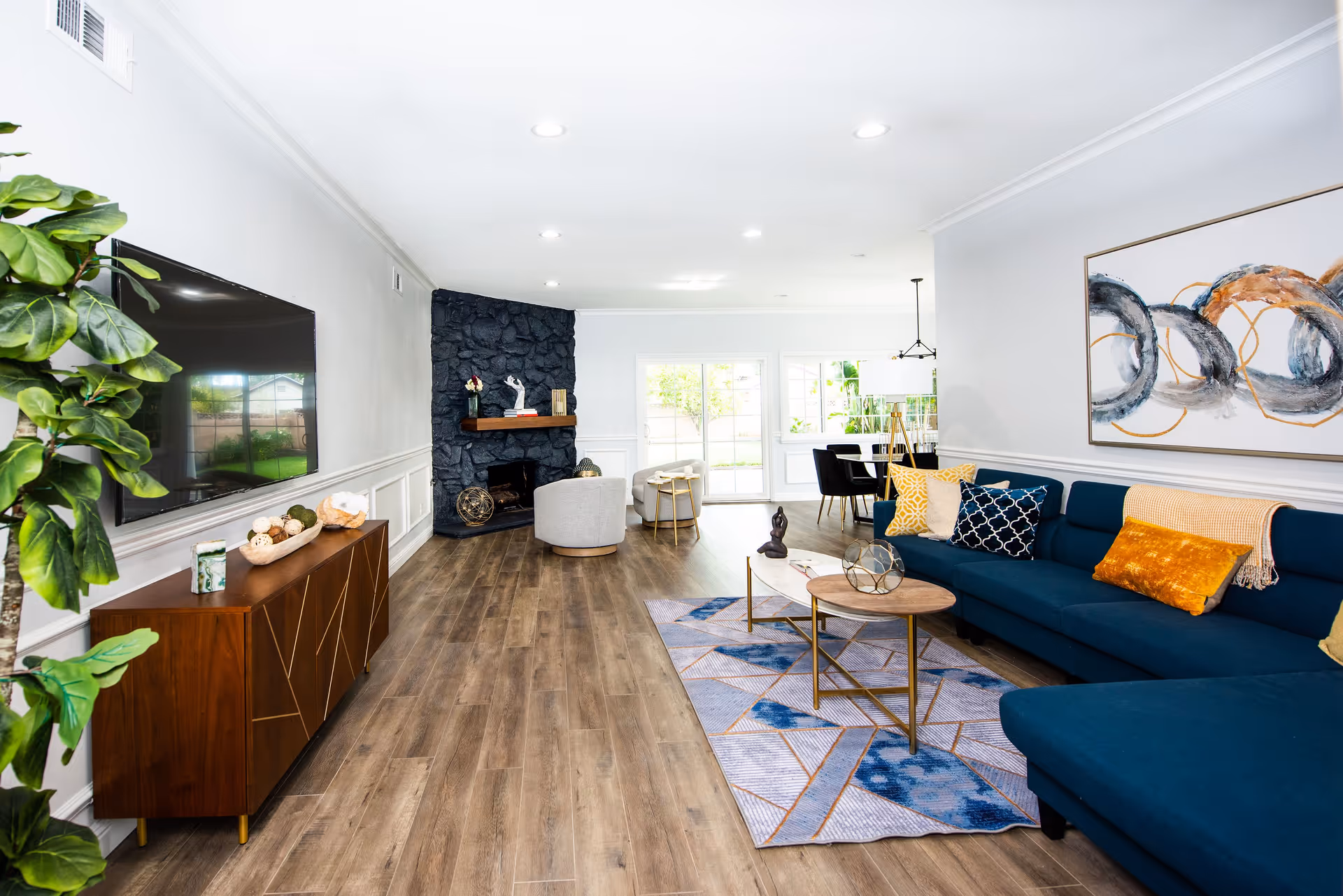 Bright modern living room with a blue sectional, coffee tables, wall-mounted TV, stone fireplace, and a dining area visible in the background.