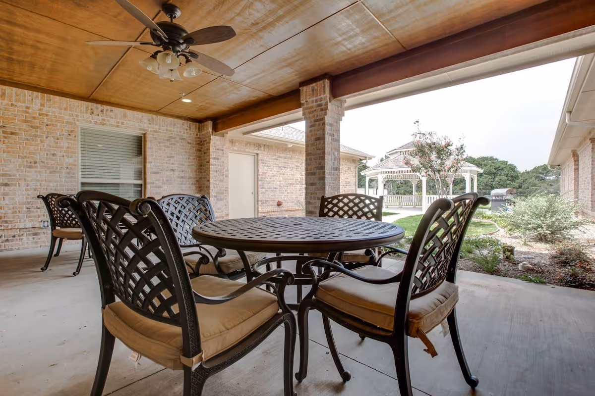 Covered outdoor patio area with a round metal table and four cushioned metal chairs. The patio has a wooden ceiling with a ceiling fan and brick walls. In the background, there is a garden area with a white gazebo, some greenery, and a barbecue grill.