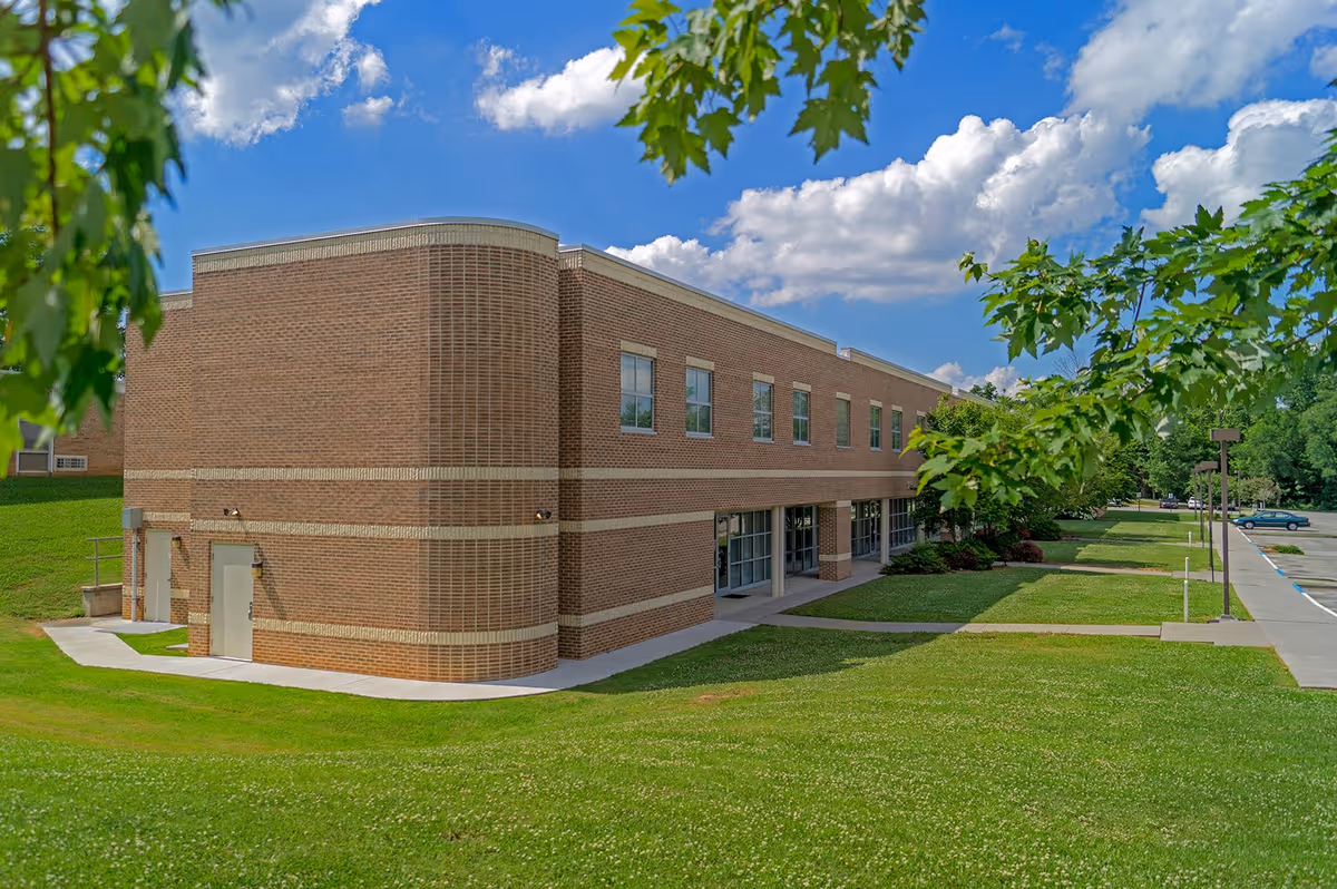 Two-story brick institutional building with a grassy lawn, trees, and a parking area under a blue sky.