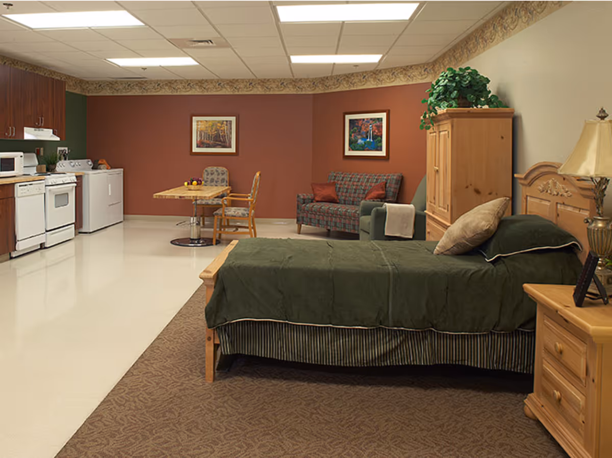 A cozy studio room in a senior living facility featuring a single bed with green bedding, a wooden nightstand with a lamp, a wooden armoire with a plant on top, a small plaid couch with two red pillows, a wooden table with two chairs, and a kitchenette area with a stove, microwave, and refrigerator. The walls are painted in warm tones with two framed pictures hanging above the couch.