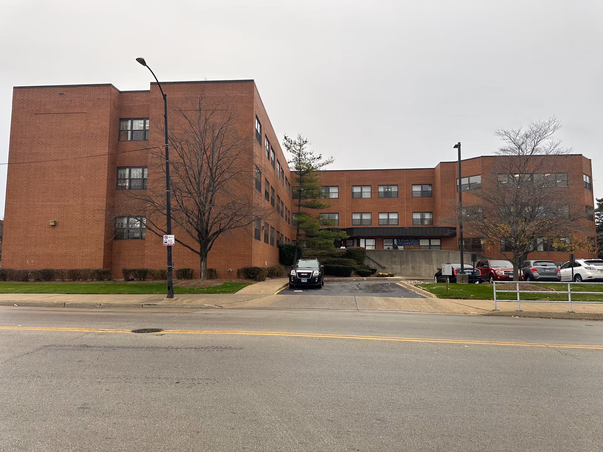 Three-story red-brick healthcare building seen from the street with a driveway, parked cars, and leafless trees.