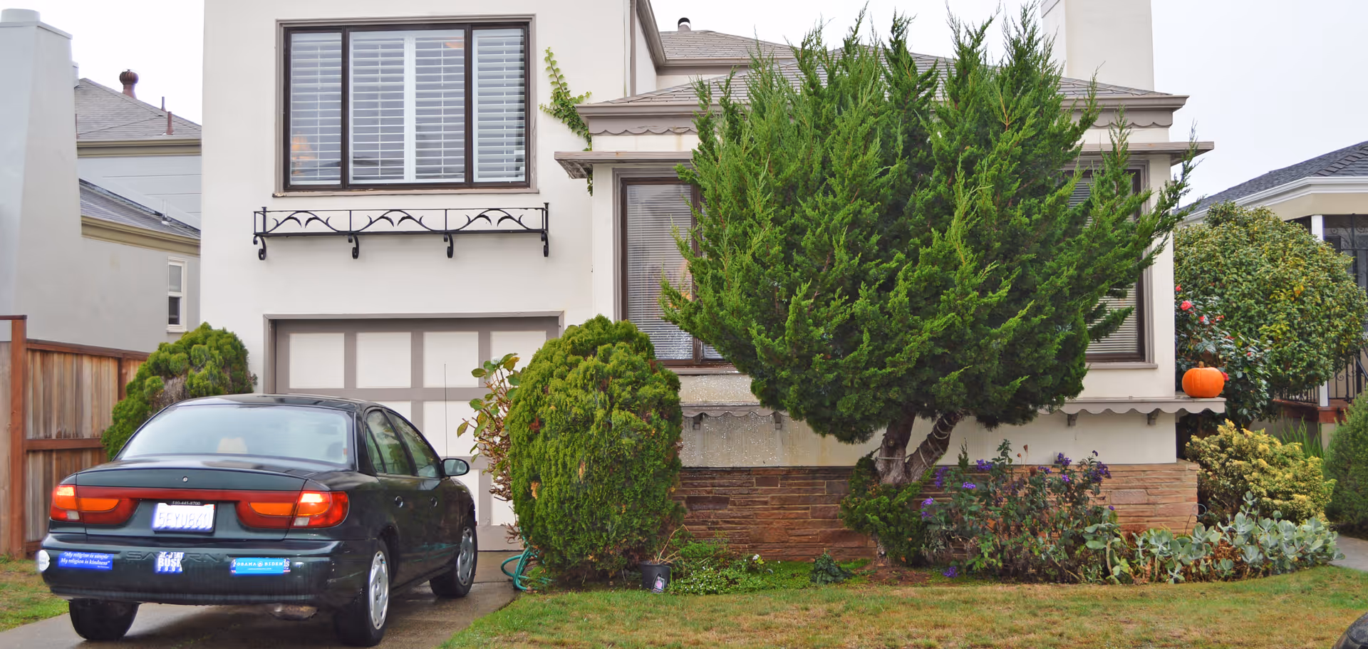 Front exterior of a house with a parked car in the driveway, a garage door, shrubs and a large tree in the front yard.