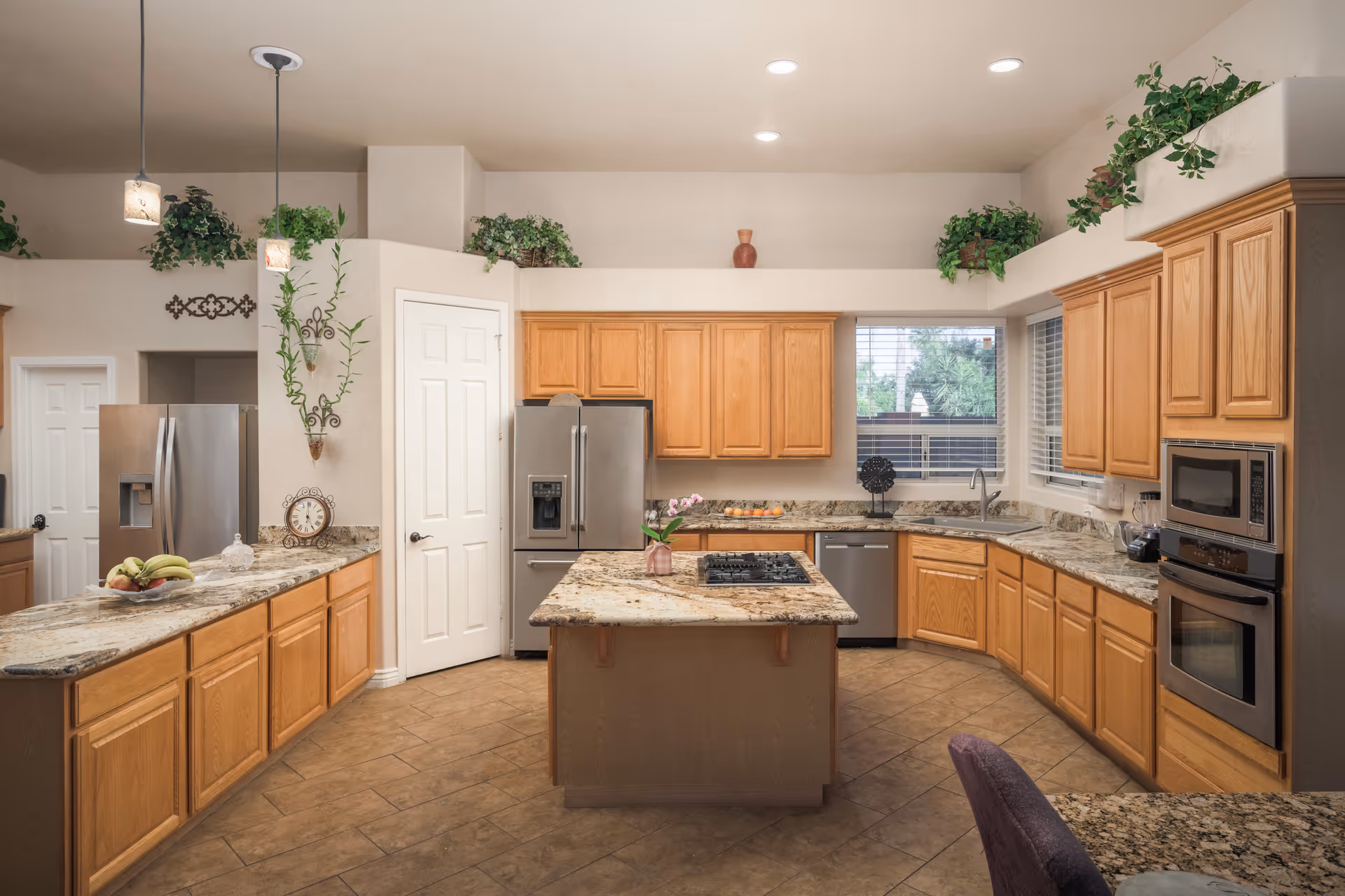 Spacious kitchen with wooden cabinets, granite countertops, a central island with a built-in stove, stainless steel refrigerator, dishwasher, microwave, and oven. The kitchen has tiled flooring, recessed ceiling lights, hanging pendant lights, and several green plants placed on top of the cabinets and walls. A window with blinds lets in natural light.