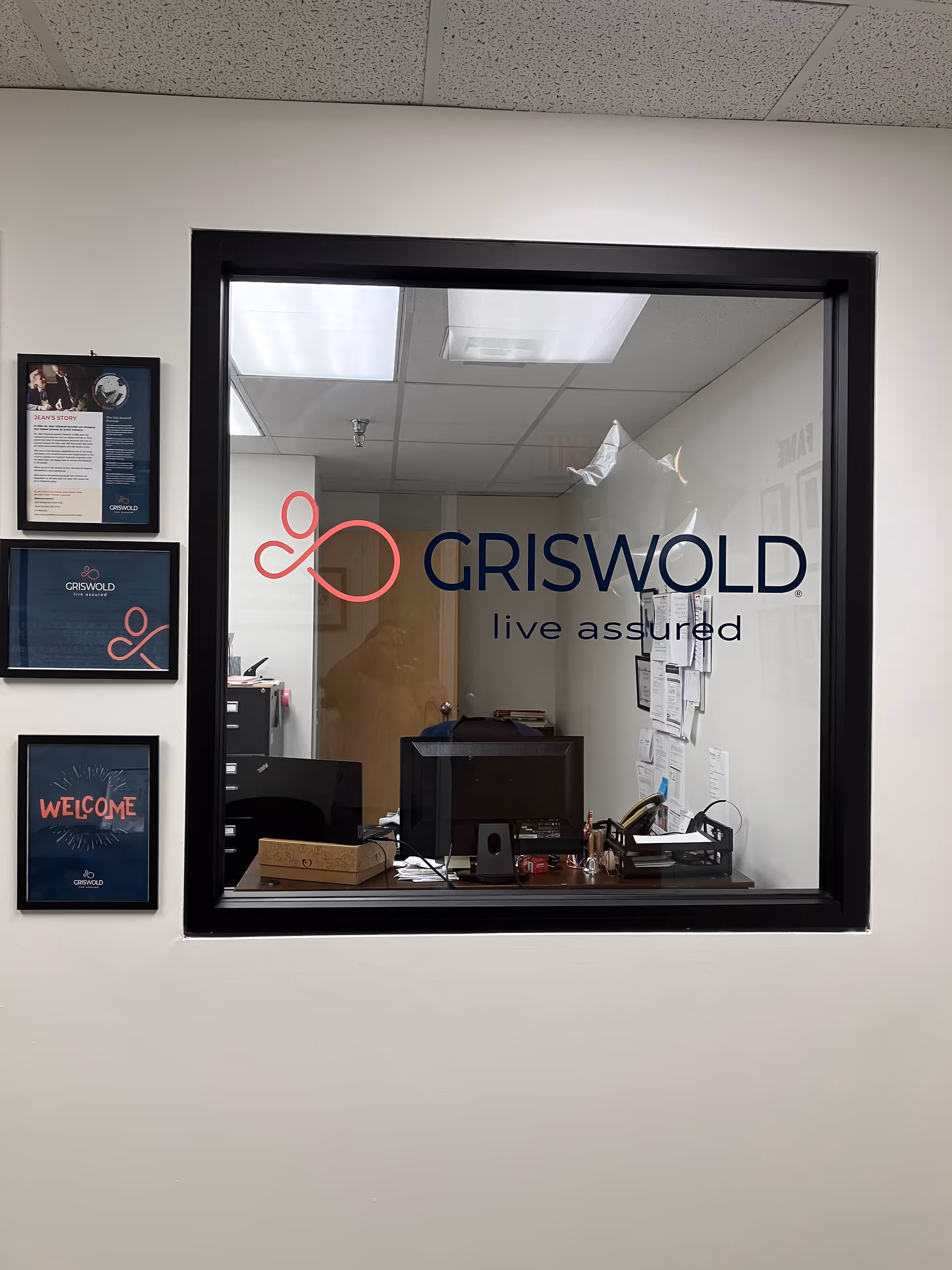 Office reception window with Griswold logo, desk, computer, and wall frames inside.