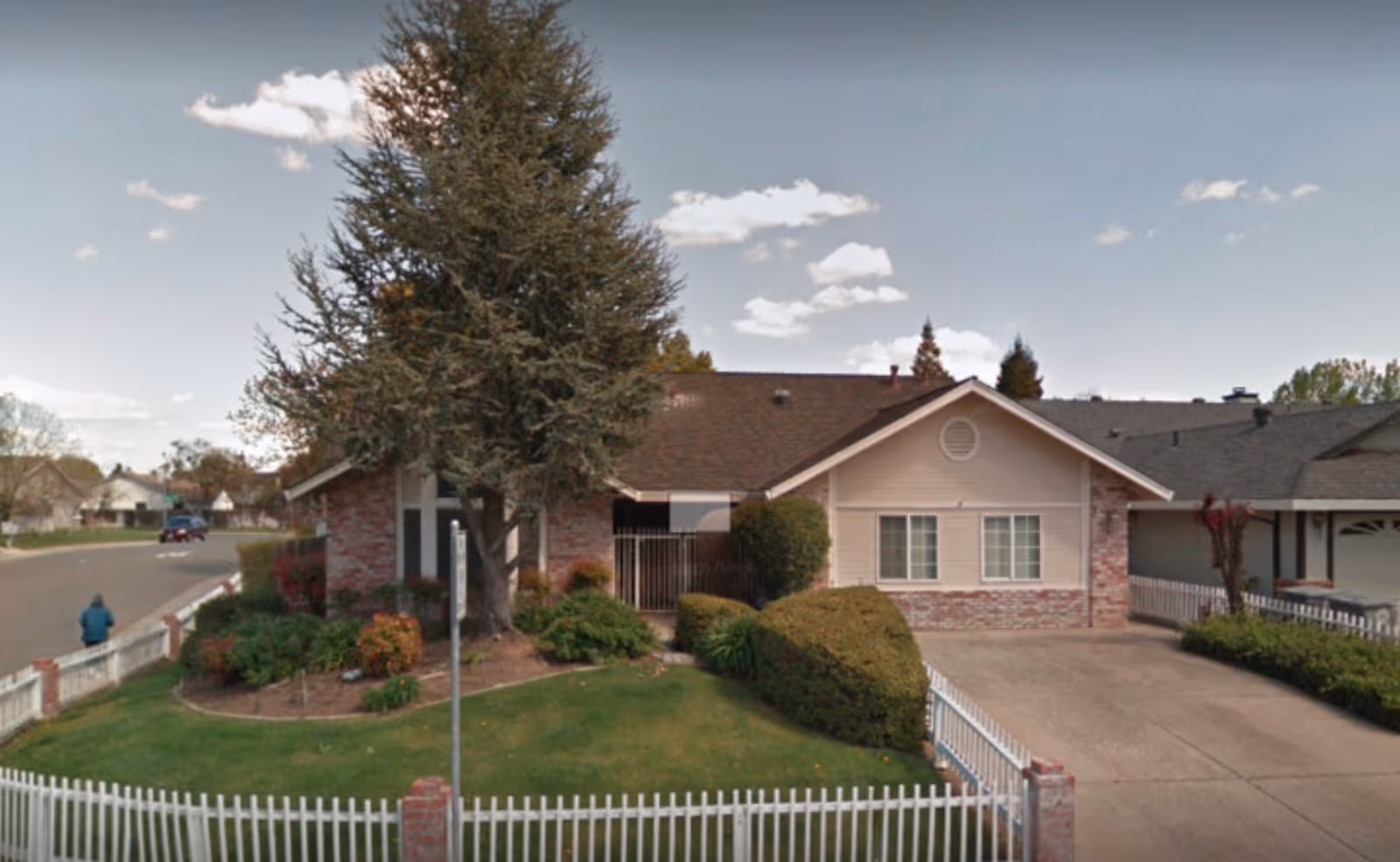 Single-story brick-and-siding residence with a large front tree, manicured lawn, white picket fence and driveway.