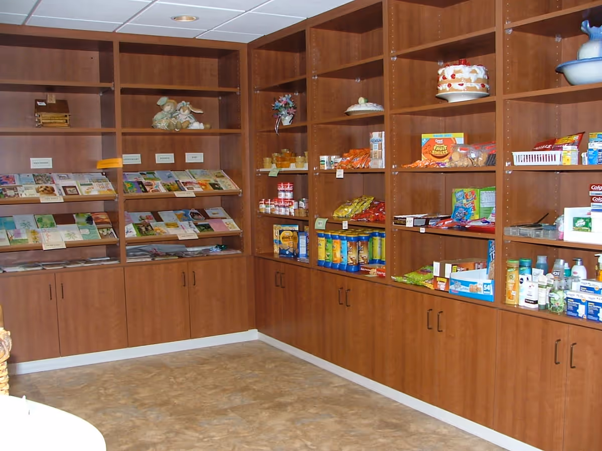 Interior view of a room with wooden shelves and cabinets filled with various items including greeting cards, canned goods, snacks, toiletries, and decorative items like a cake model and stuffed animals.