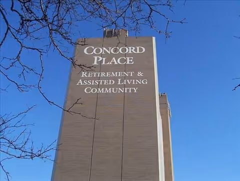 Tall brick building facade with large signage reading "Concord Place Retirement & Assisted Living Community" against a clear blue sky.