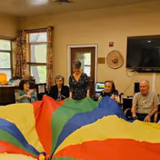 A group of five elderly people sitting in a circle indoors, holding the edges of a colorful parachute with red, yellow, green, and blue sections. The room has windows with curtains, a wall-mounted TV, a hat hanging on the wall, and a door in the background.