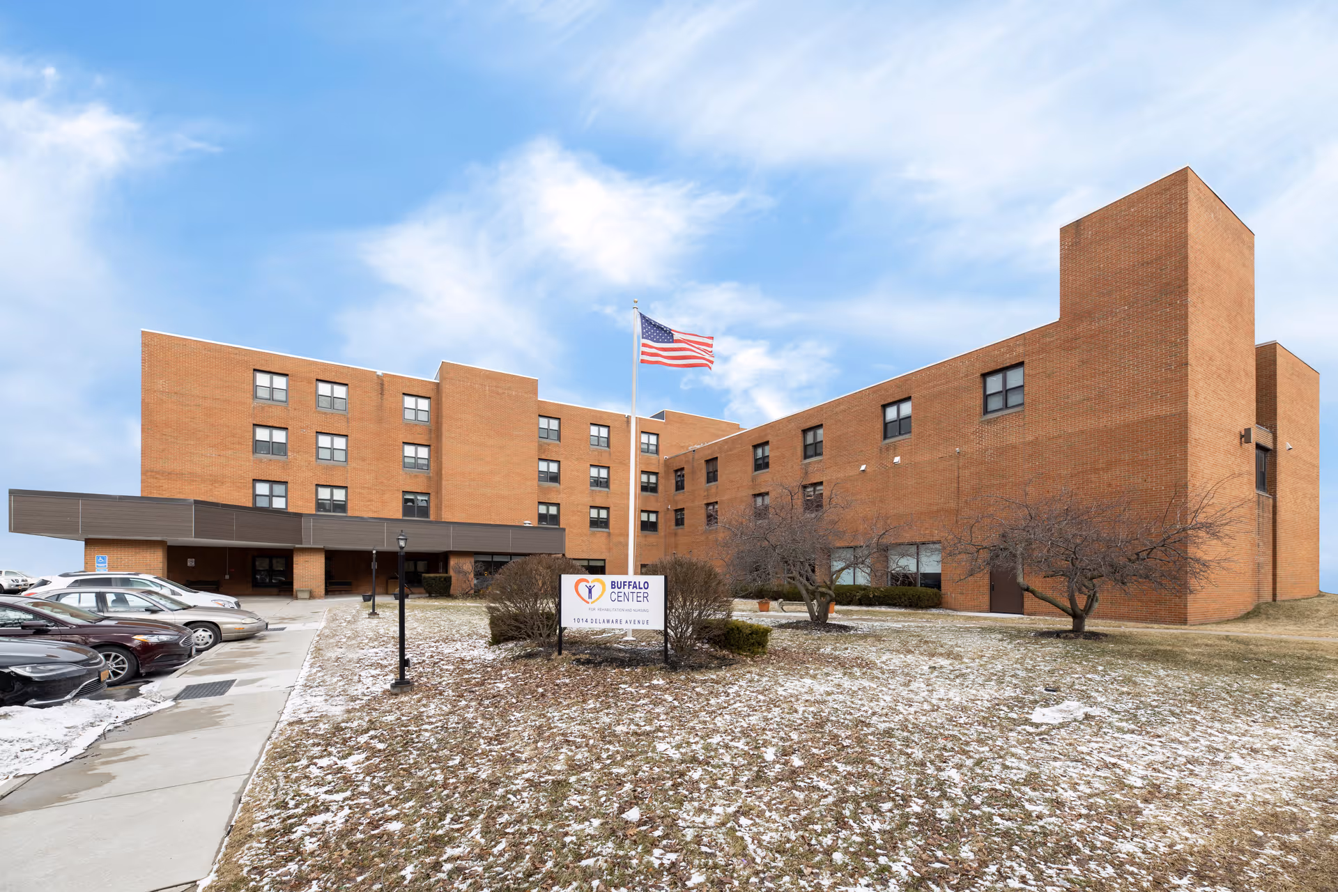 Exterior view of a multi-story brick building with an American flag on a flagpole in front. There is a sign that reads 'Buffalo Center' along with the address 1034 Delaware Avenue. Several cars are parked along a driveway on the left side, and the ground is lightly covered with snow patches under a partly cloudy sky.