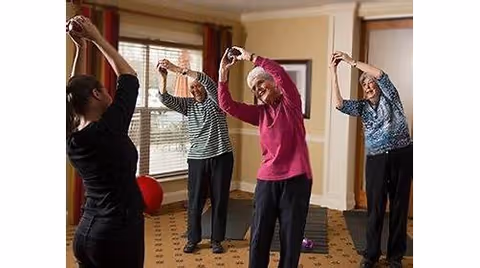 A group of three elderly women stretching their arms overhead while following an instructor in a room with large windows and carpeted floor.