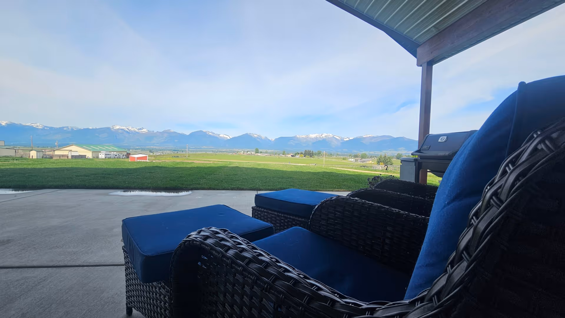 Outdoor patio area with wicker chairs and blue cushions under a covered roof, overlooking a green lawn and distant snow-capped mountains under a clear sky.
