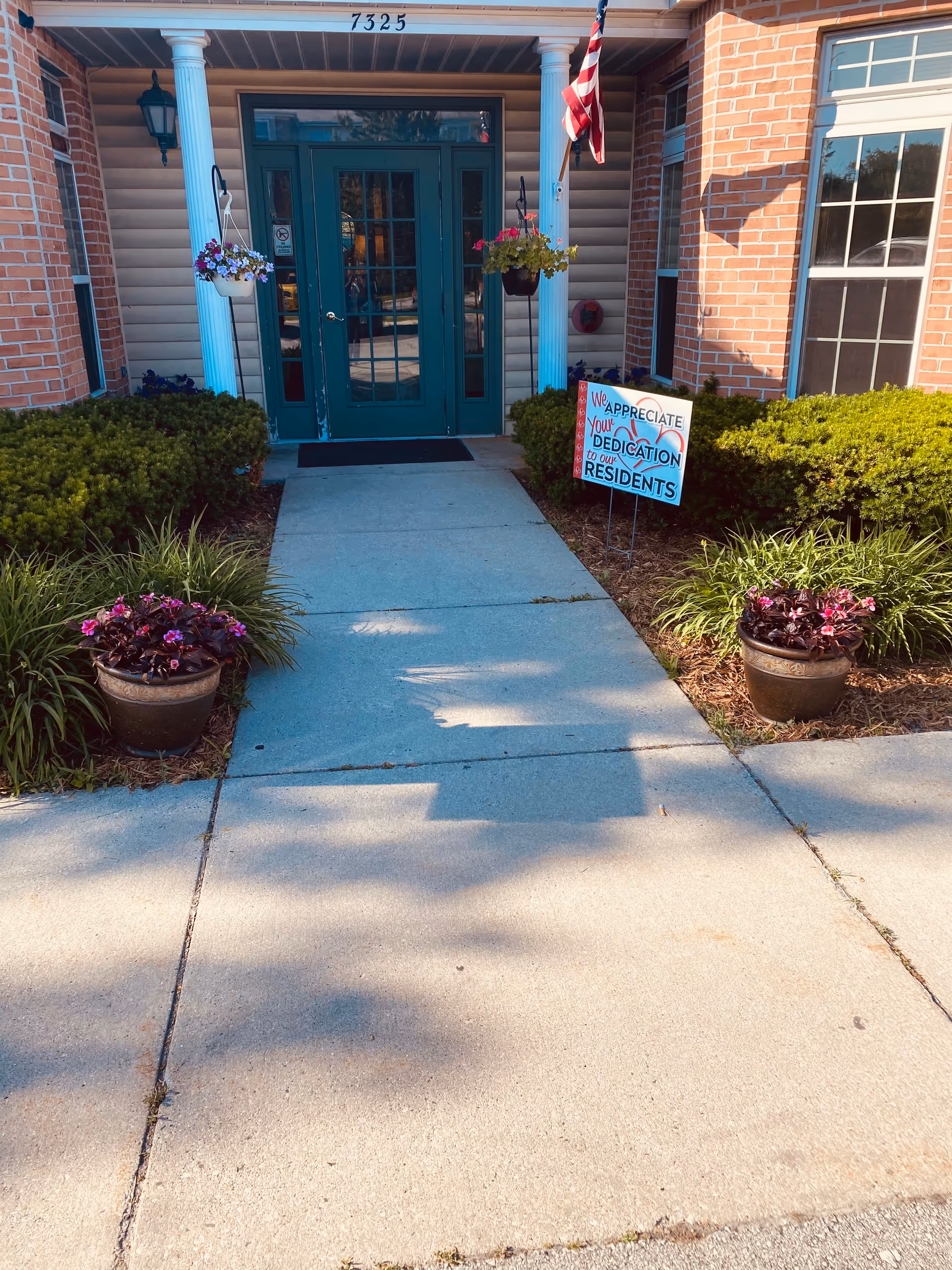 Entrance to a building with green double doors, white columns, and brick walls. There are hanging flower pots on either side of the doors and two large flower pots with purple flowers on the ground. A sign on the right side of the walkway reads 'We appreciate your dedication to our residents.' The building number 7325 is displayed above the entrance, and an American flag is mounted on the right column.