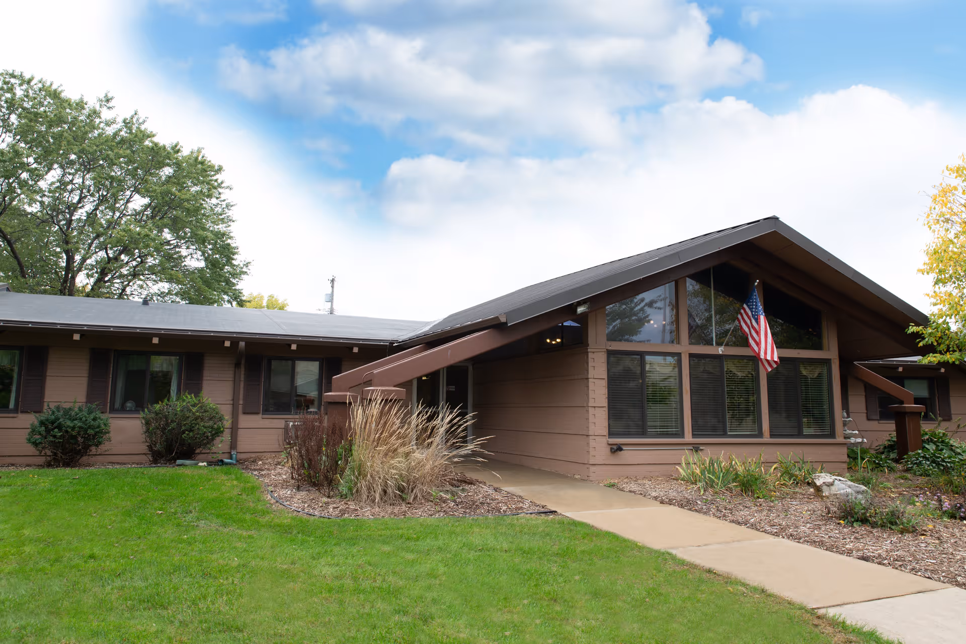 Exterior view of a single-story brown building with large windows and an American flag mounted near the entrance. The building is surrounded by green grass, shrubs, and trees under a partly cloudy sky.