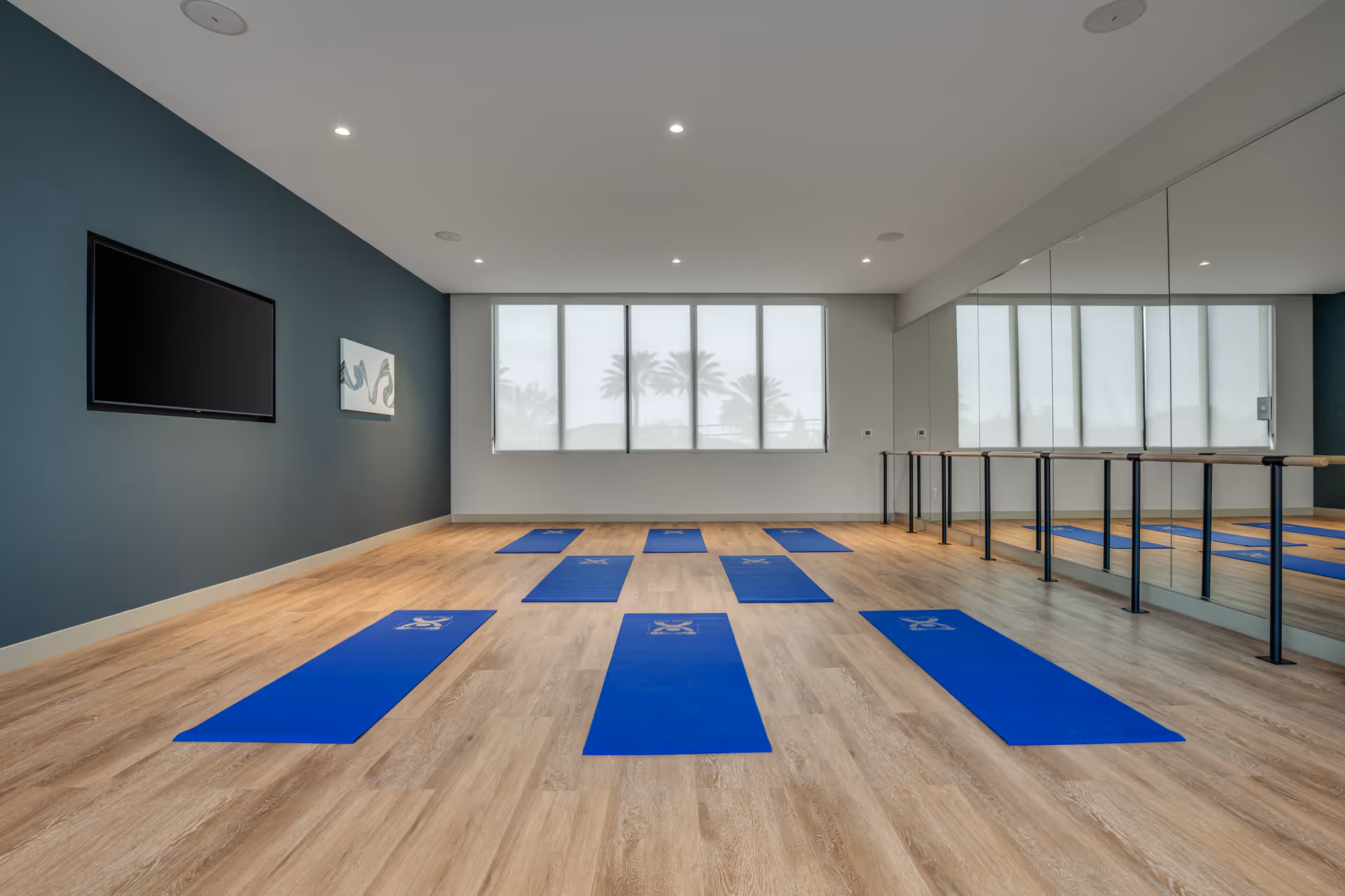 A spacious exercise or yoga room with wooden flooring and blue yoga mats neatly arranged on the floor. The room has a large window with blinds, a wall-mounted TV, a piece of abstract wall art, and a mirrored wall with ballet barres. The walls are painted in a combination of teal and white.