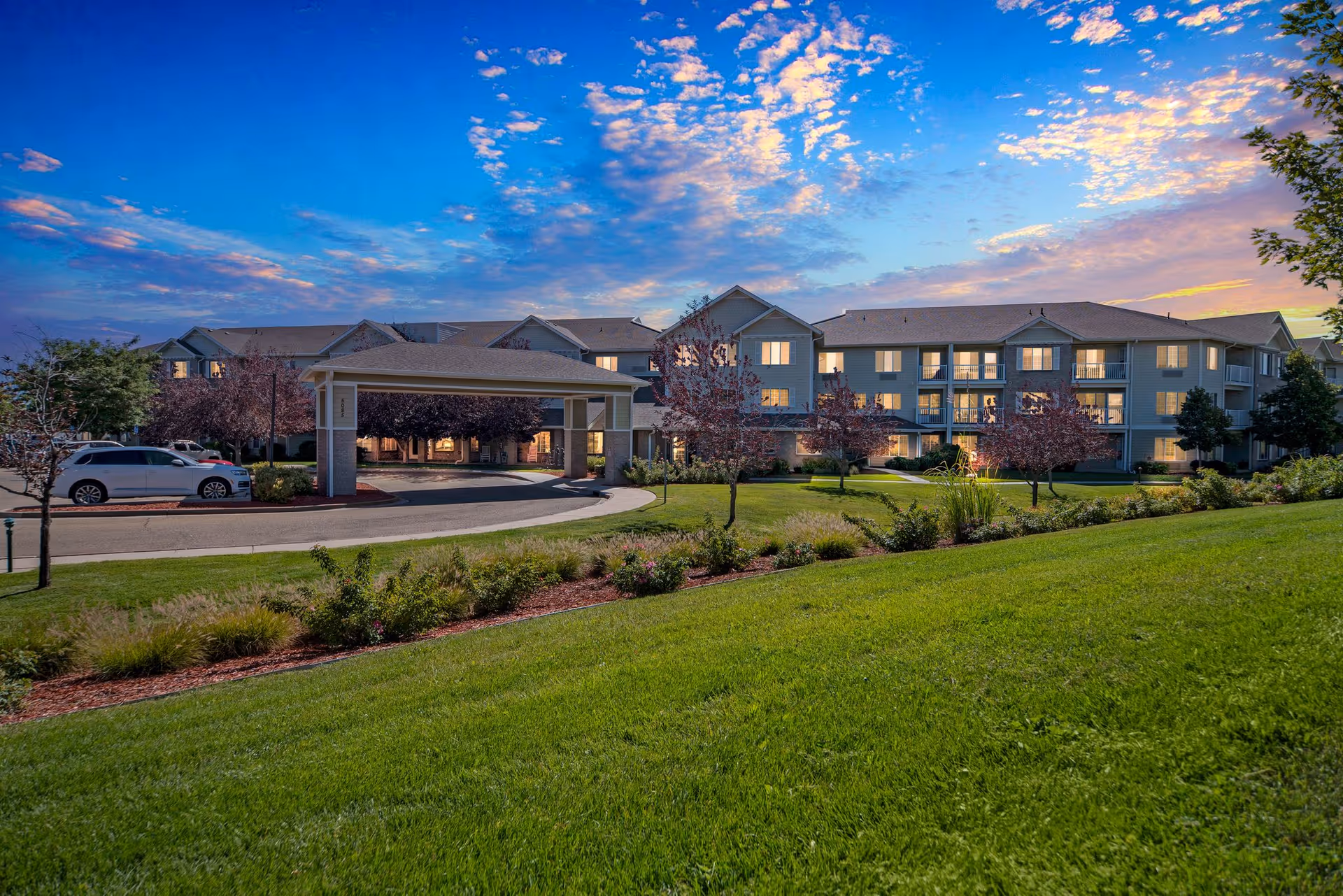 Well-lit three-story senior living building with a covered entrance, parked cars, and manicured lawn under a colorful evening sky.