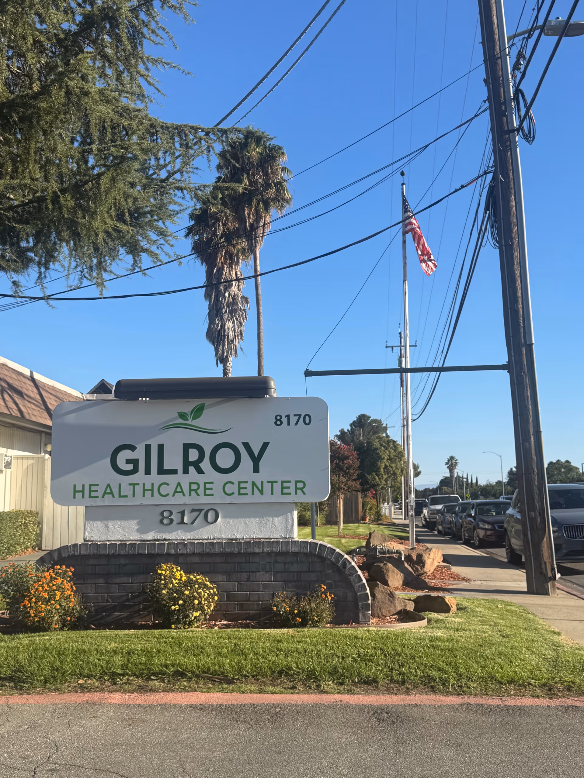 Exterior view of Gilroy Healthcare Center sign with street, trees, and cars.