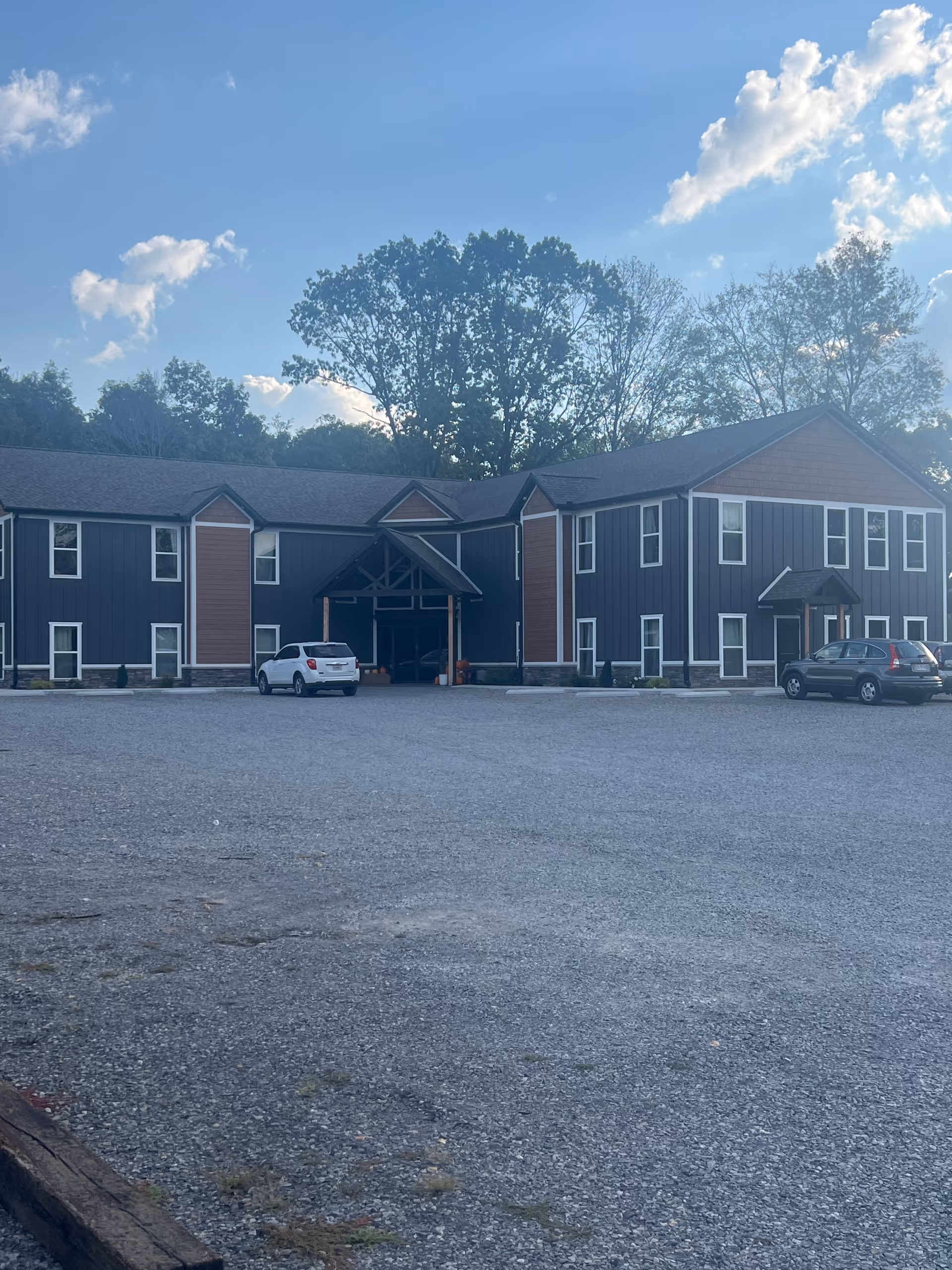 Front view of a blue and brown senior living facility with a gravel parking lot.
