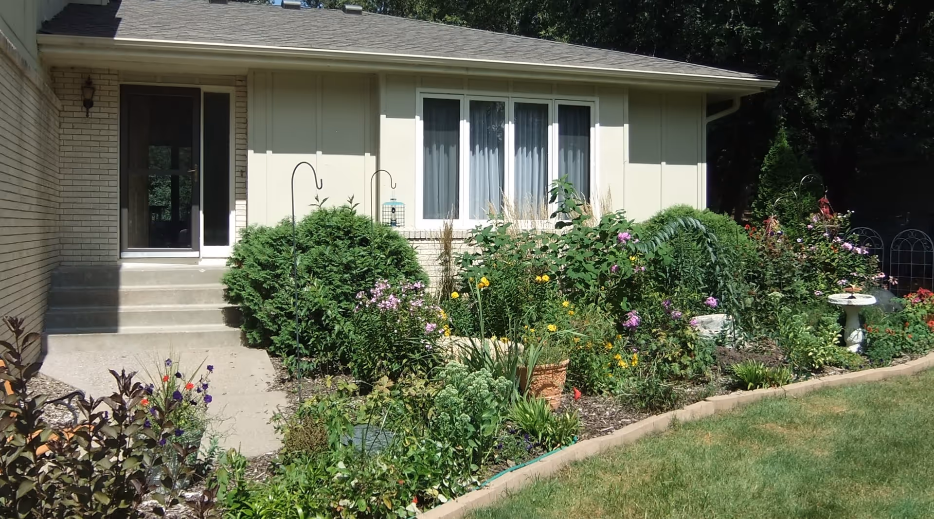 Front exterior view of a single-story building with a garden in front. The garden is filled with various green plants and colorful flowers. There is a concrete pathway leading to a door with steps, and a window with white curtains is visible on the building's wall.