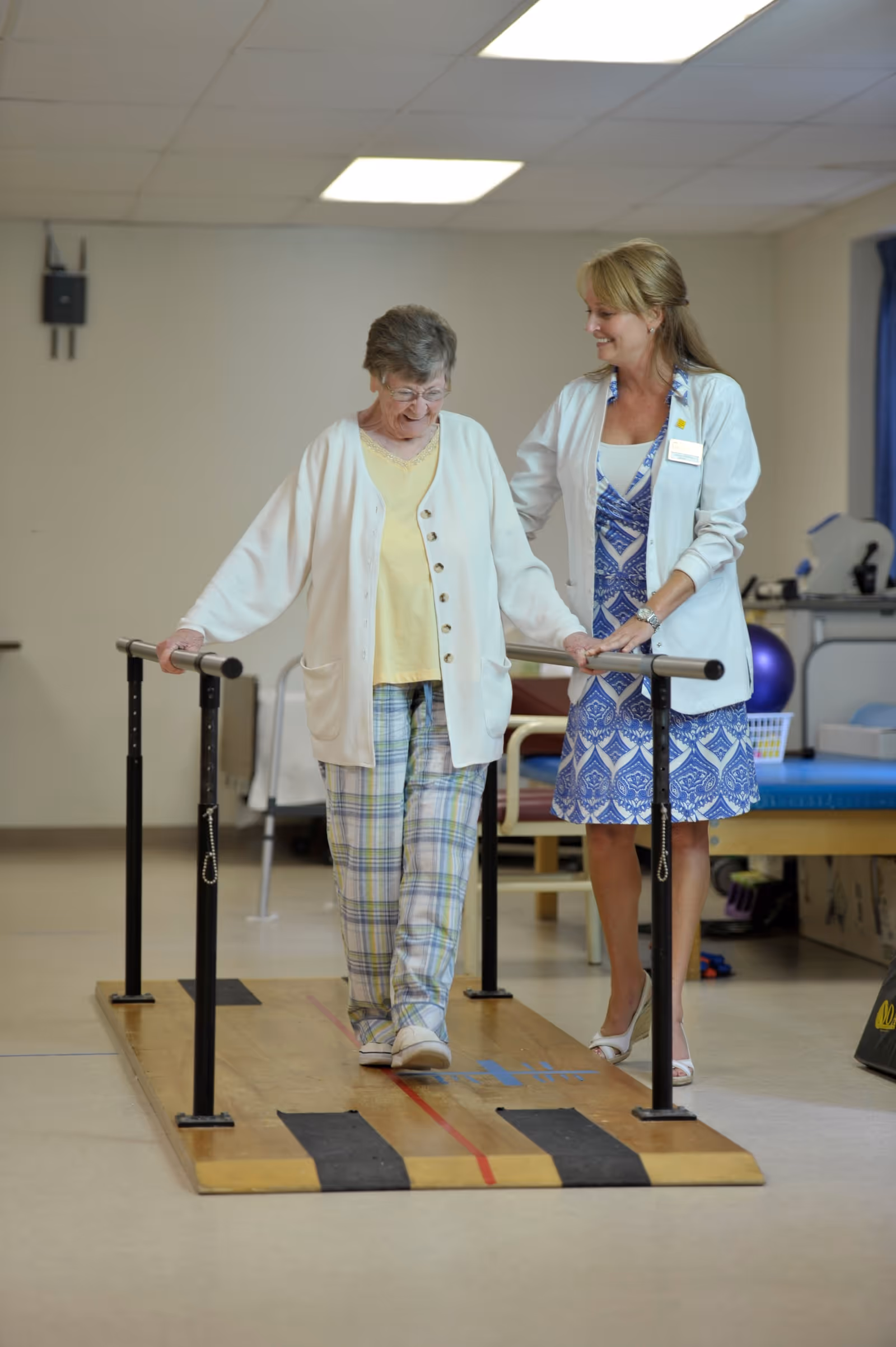 An elderly woman using parallel bars for physical therapy while being assisted by a female healthcare professional in a rehabilitation center room.
