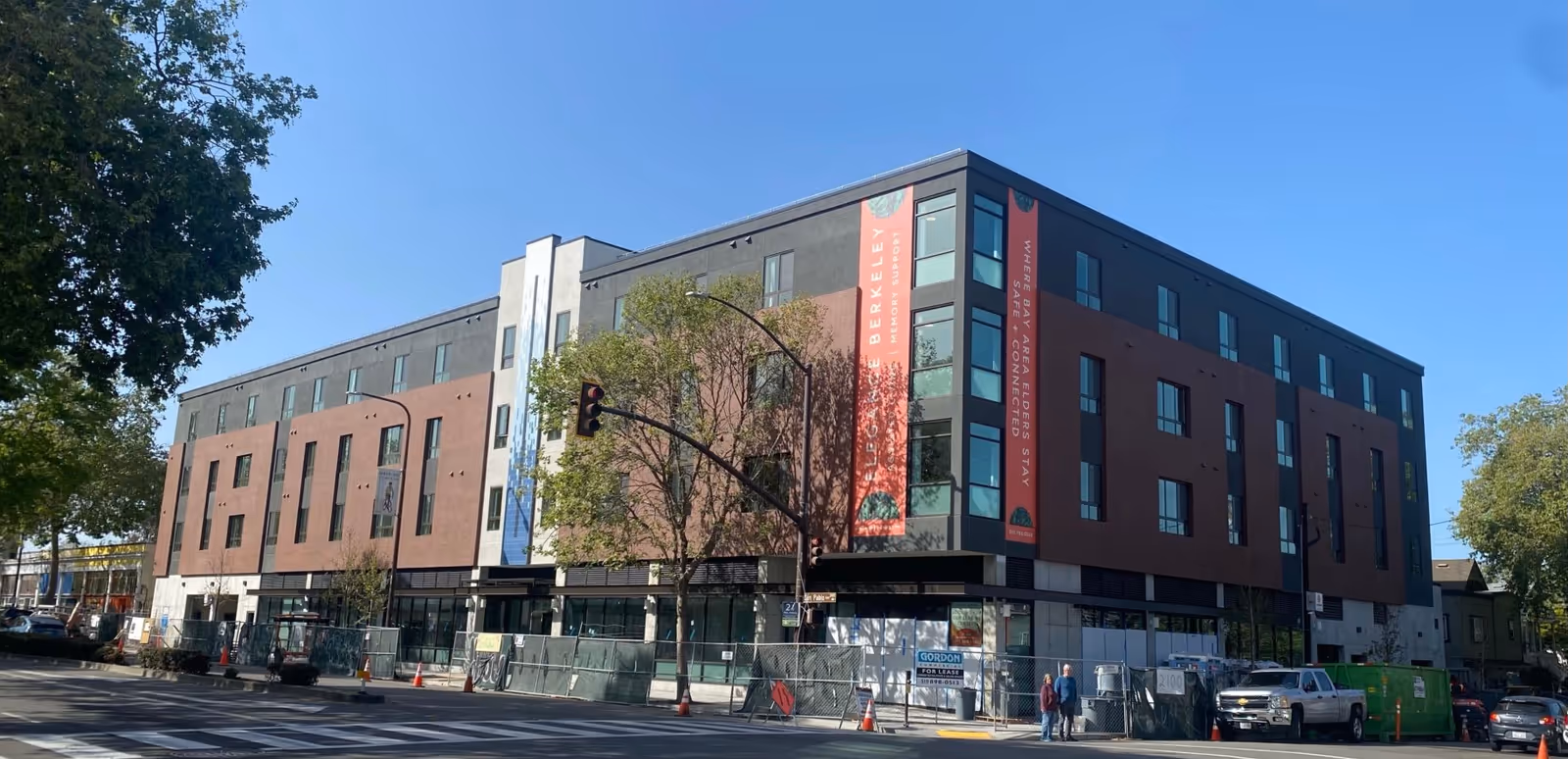 Modern four-story Elegance Berkeley senior living building with banners, trees, fencing, and blue sky