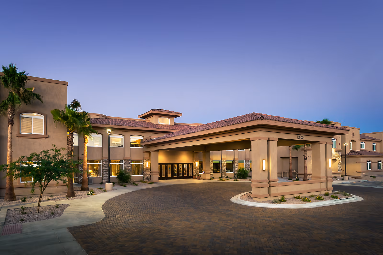 Exterior view of MorningStar Assisted Living & Memory Care of Fountain Hills building at dusk, featuring a covered entrance with pillars, palm trees, and well-lit windows.