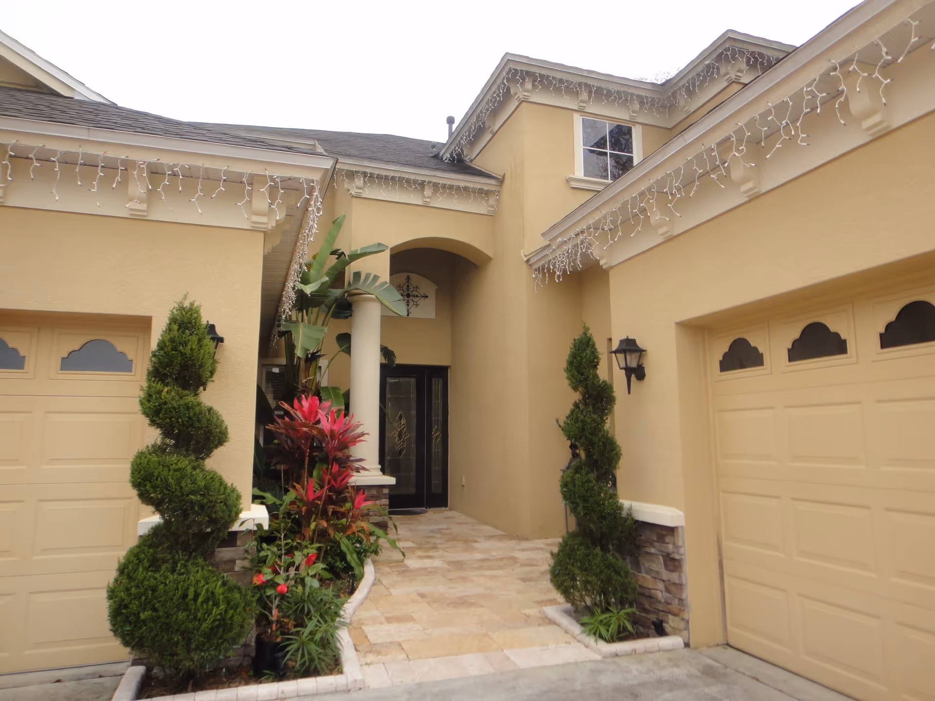 Front entrance of a beige stucco building with two garages on either side, decorative spiral-shaped topiary plants, red and green foliage, and a tiled walkway leading to a double glass door under an archway. White string lights hang along the roof edges.