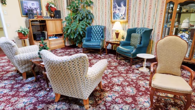 Cozy senior living sitting room with upholstered armchairs, side tables, plants and patterned carpet.