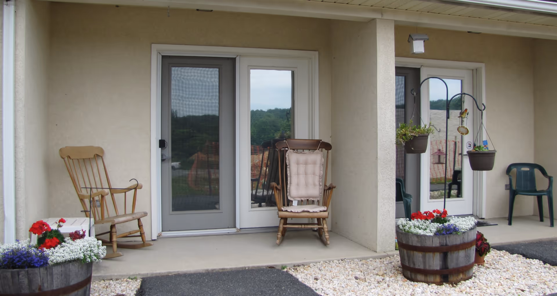 Covered porch of a senior living unit with two rocking chairs, potted flower barrels, hanging plants, and glass doors.