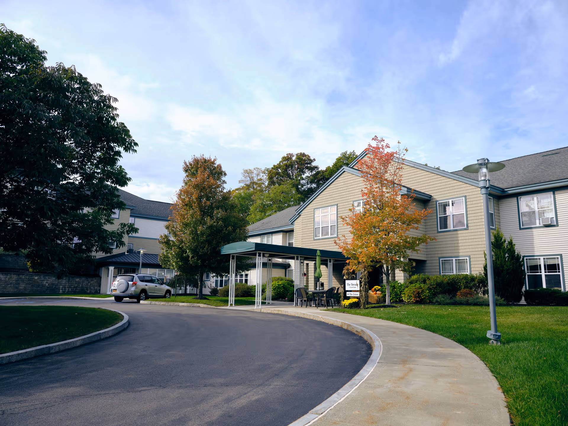 Exterior view of a senior living facility named Terrace At Glen Eddy with a curved driveway, green lawn, trees with autumn foliage, and a covered entrance with chairs and a table.