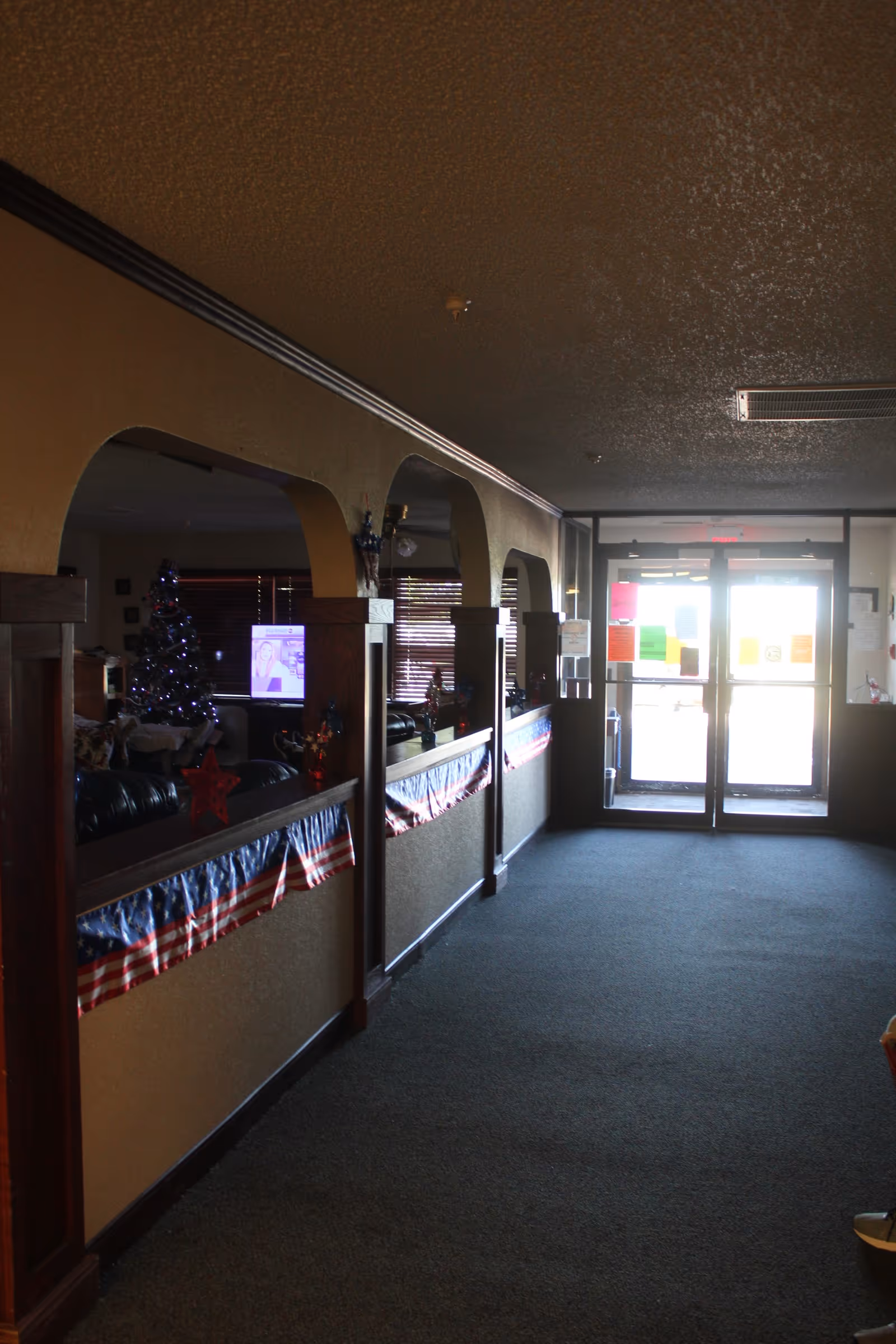 Interior hallway of a facility with carpeted floor and beige walls featuring wooden trim and arches. The left side shows a living area decorated with a Christmas tree and American flag bunting along the half walls. At the end of the hallway, there is a glass double door entrance with various colorful notices posted on the glass.