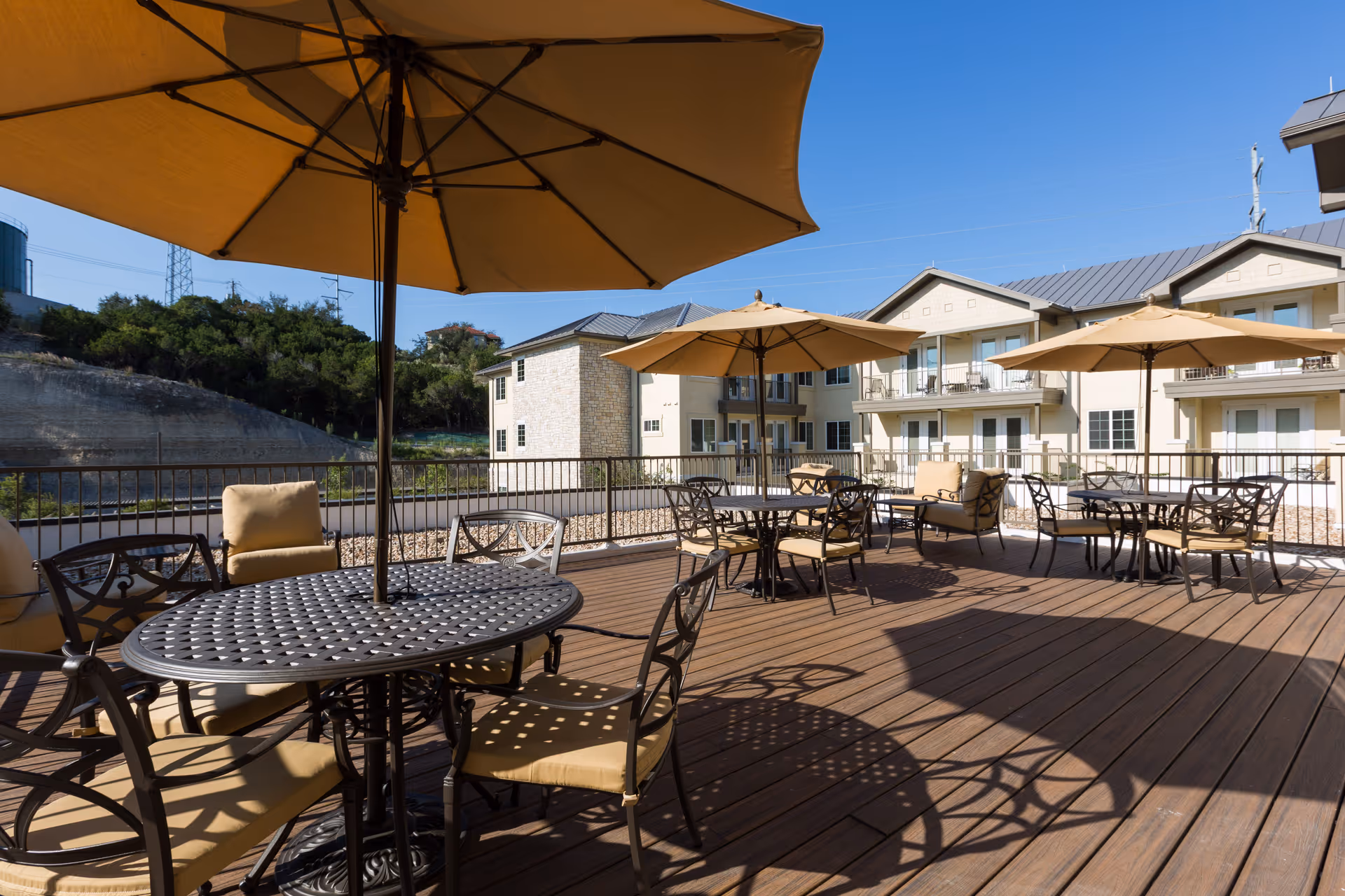 Outdoor patio area with multiple round metal tables and cushioned chairs under large beige umbrellas, with a view of a multi-story residential building and a hillside in the background under a clear blue sky.