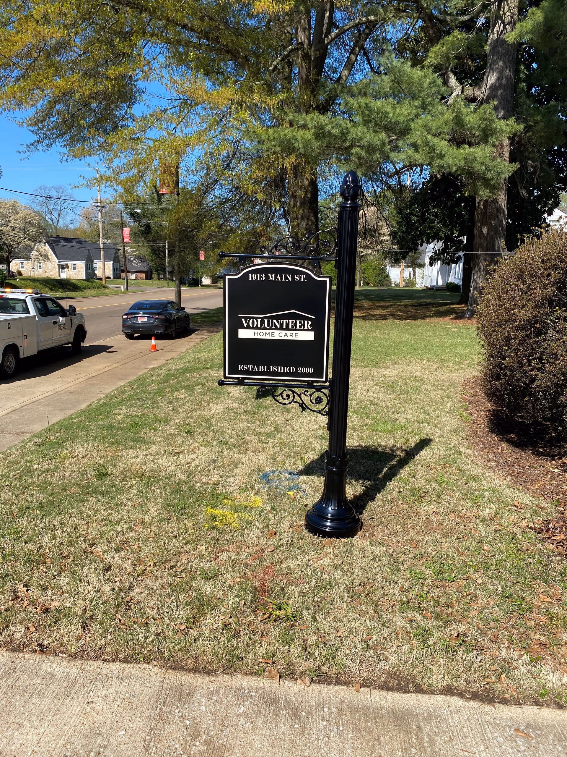 A black decorative sign for Volunteer Home Care stands on a grassy lawn beside a sidewalk.