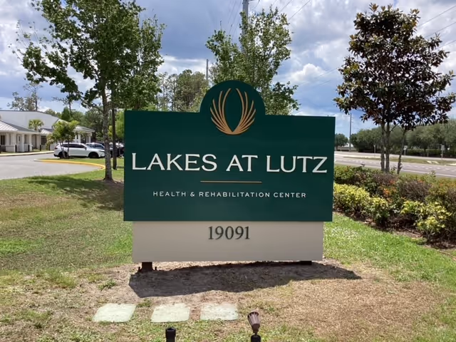 Exterior sign reading Lakes at Lutz Health & Rehabilitation Center with surrounding trees and landscaping.