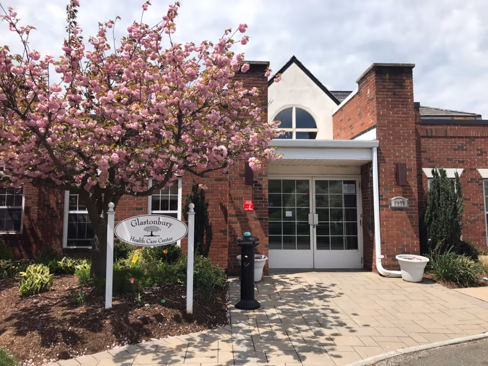 Brick-front entrance of the Glastonbury Health Care Center with double doors, a flowering pink tree, and a sign in the landscaped front.