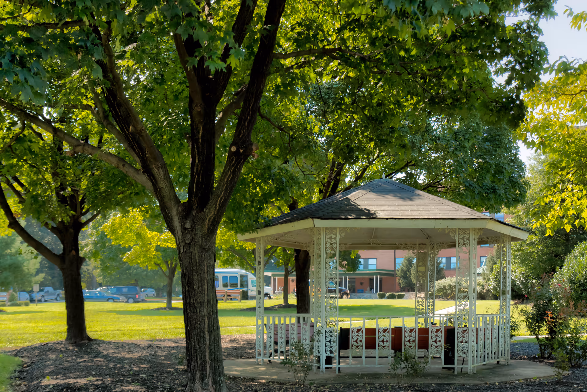 A white gazebo with decorative metal railings and a shingled roof sits in a green park-like area surrounded by large leafy trees. In the background, there is a brick building and several parked vehicles visible through the trees.