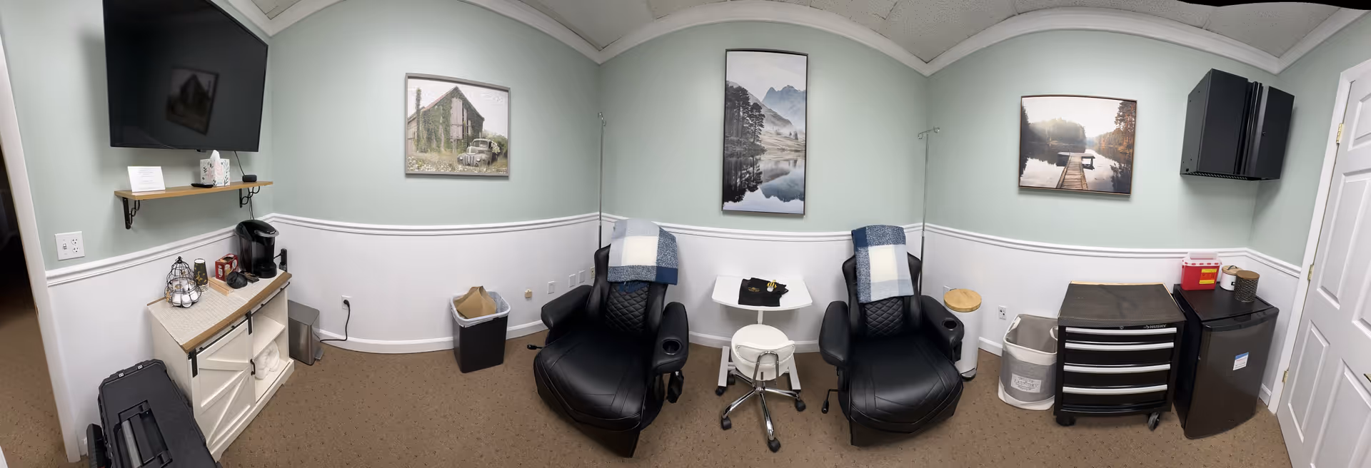 Well-lit treatment room with two reclining chairs, blankets, wall art, and a coffee station.