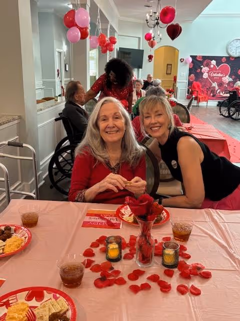 Two women smiling and sitting at a table decorated with red rose petals, candles, and Valentine's Day themed items. The table has plates with snacks and drinks. In the background, other people are seated and Valentine decorations including balloons and a banner are visible.