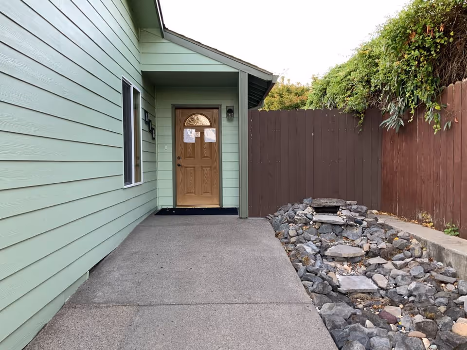 Green-siding senior home entrance with wooden door and rocky landscaping along a path.