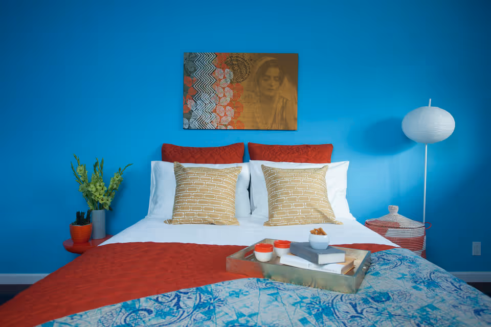 A brightly colored bedroom with a neatly made bed against a turquoise wall, patterned pillows and bedding, a piece of wall art above, and bedside decor including a lamp and plants.