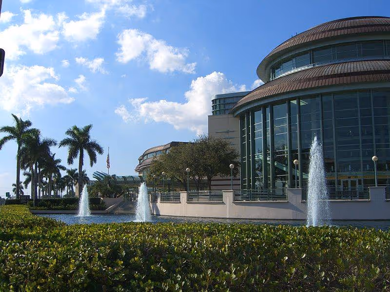Exterior view of a curved-roof glass building with fountains and palm trees.