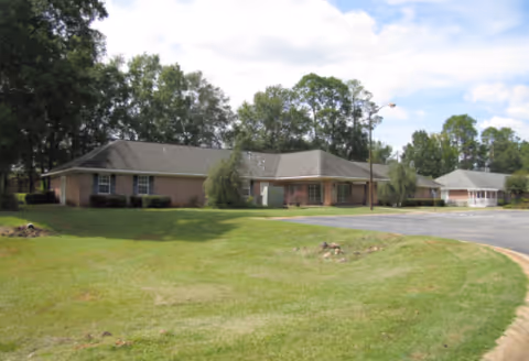 Single-story brick senior living facility with manicured lawn, trees, and curved driveway.