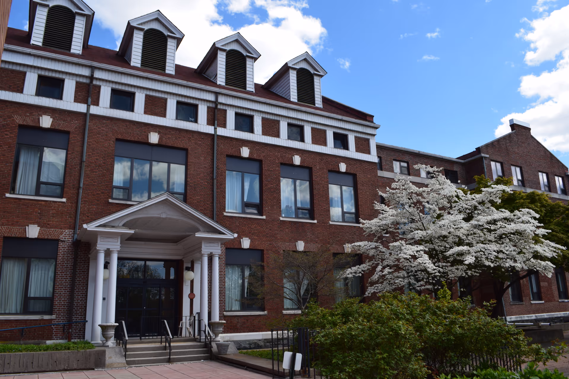 Exterior view of a multi-story brick building with white trim and dormer windows on the roof. The entrance features a small portico with columns and steps leading up to glass doors. There are trees and bushes with white blossoms in front of the building under a partly cloudy blue sky.