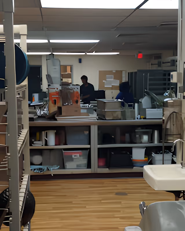 Interior view of a facility kitchen/prep area with shelving, storage bins, countertop equipment, a sink in the foreground, and staff working in the background.