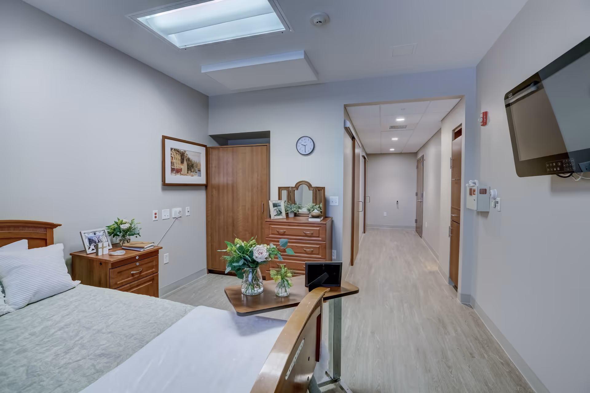 Senior living room with bed, nightstands, dresser, plants, and a hallway leading to doors.