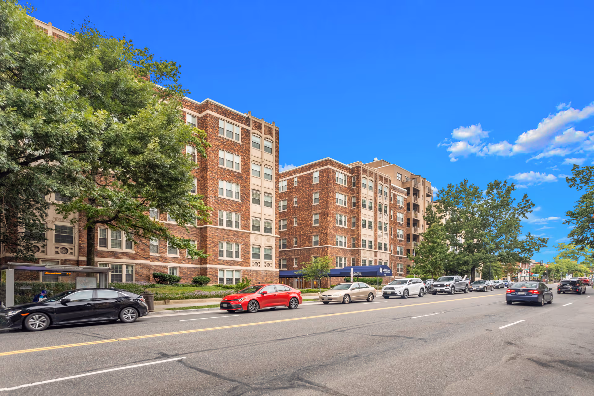 Street view of a multi-story brick building with several windows and a blue awning entrance, surrounded by trees and parked cars along the road under a clear blue sky.
